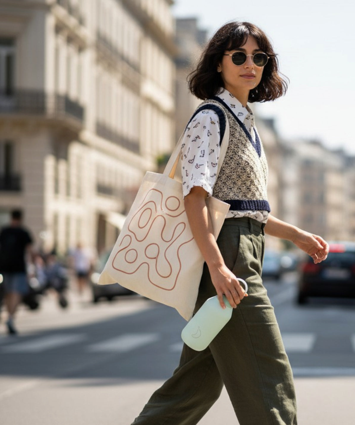 Mujer con gafas de sol caminando por la calle, con estilo urbano, llevando un bolso de tela y una botella Chilly's Series 2 polen 350ml en la mano.